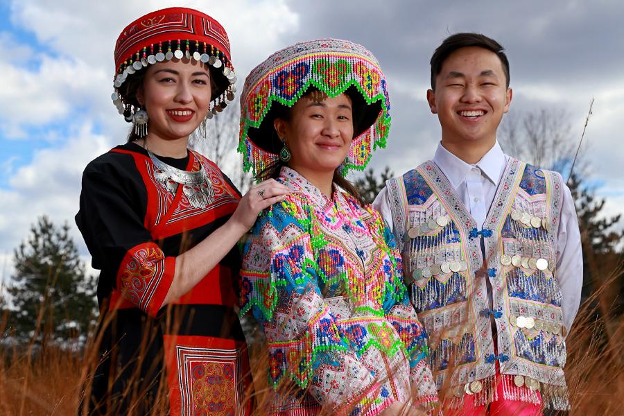 Three Asian students model traditional Hmong clothing, jewelry and story cloth.