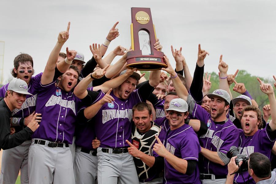 Men's baseball cheers and holds up an NCAA trophy on a baseball diamond.